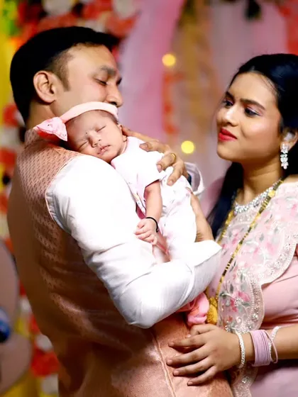 A tender family portrait from a naming ceremony. The father holds their sleeping newborn while the mother looks on with love, creating a warm and intimate photograph.