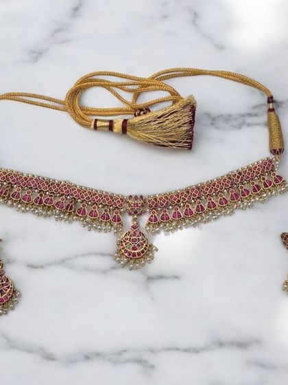 A delicate and stunning choker set. This gold-plated silver necklace with matching earrings features intricate work with pink stones and tiny pearl drops, complete with an adjustable tassel tie.