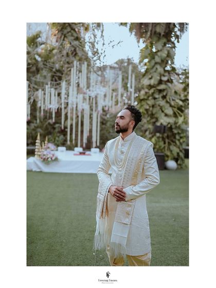 The groom looking dapper in his white sherwani, standing in a garden with beautiful hanging decorations.
