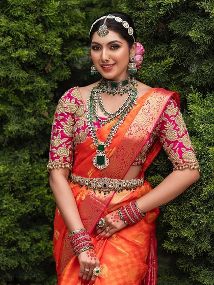 A smiling bride surrounded by lush greenery. This outdoor portrait showcases a vibrant Kanjeevaram saree and a full set of diamond and emerald jewellery.