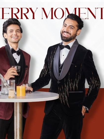 Merry moments. Two men in stylish tuxedos, one in maroon velvet and the other in black with gold embroidery, sharing a laugh. Our suits are made for celebrations.