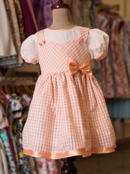 A sweet gingham dress in a lovely peach color. The puff sleeves and simple bow create a look of classic, storybook charm.