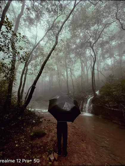A person stands with an umbrella in a dense, foggy forest in Matheran, with a small waterfall nearby. This image is full of atmosphere, capturing the feeling of being immersed in nature during the monsoon.