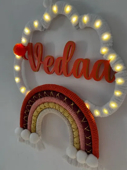 The 'Vedaa' name hanging glowing beautifully with fairy lights. This piece uses a warm, sunset-inspired color palette for the rainbow.