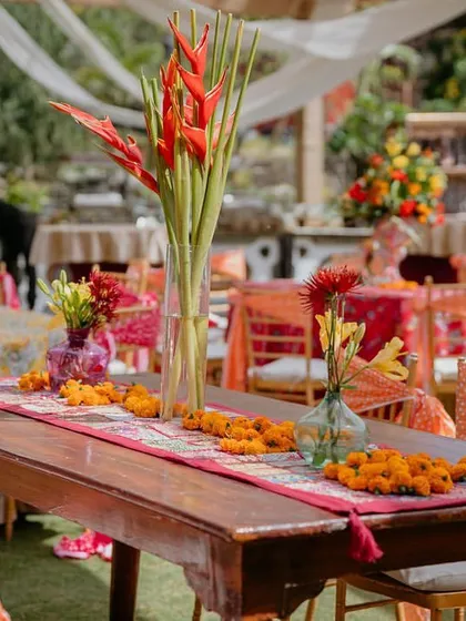 A dining table setup from the 'Rajasthani Folklore' Mehendi, featuring a traditional runner and bright red heliconia flowers in a tall vase.