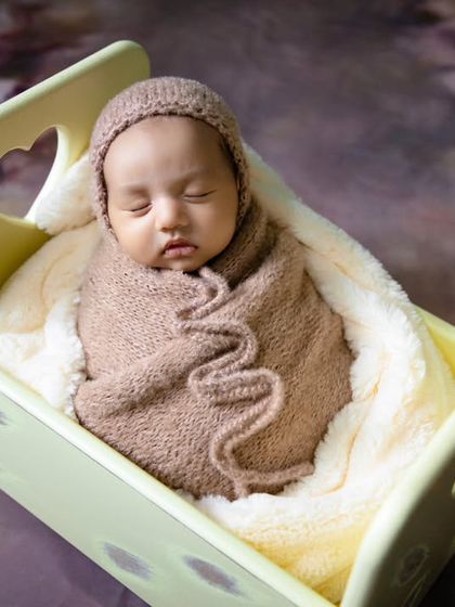 A cozy shot of a baby nestled in a miniature bed prop, with a textured brown wrap and bonnet.