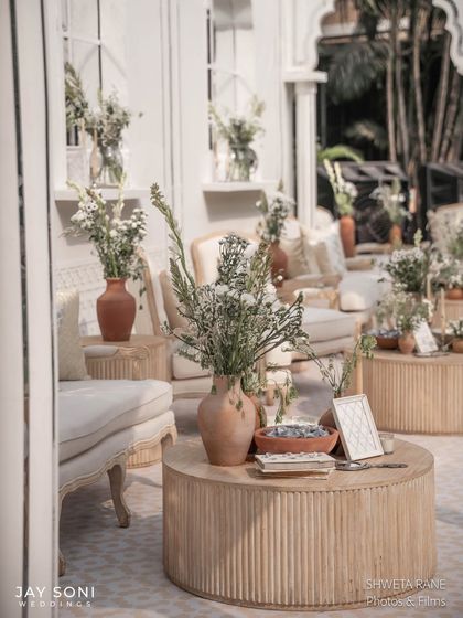A close-up of a seating group within the ivory-themed lounge, featuring a modern slatted wood coffee table and rustic terracotta vases with delicate white wildflowers.