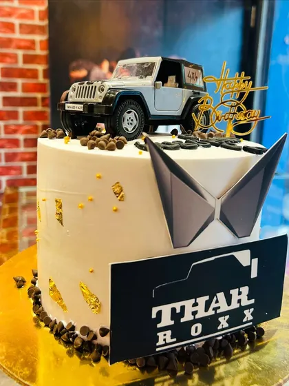 A white Mahindra Thar-themed cake with gold leaf accents and a "Thar Roxx" plaque.