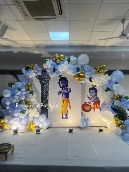 A grand and traditional Krishna-themed decoration for a naming ceremony or birthday. The stage features cutouts of Little Krishna against backdrop panels, framed by blue, white, and gold balloon arches and a beautiful peacock prop.