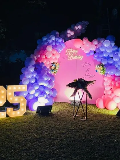 An outdoor 25th birthday setup on a lawn, featuring a pink backdrop with a balloon arch. The decoration is completed with a 'Happy Birthday' neon sign and large, illuminated '25' marquee numbers.