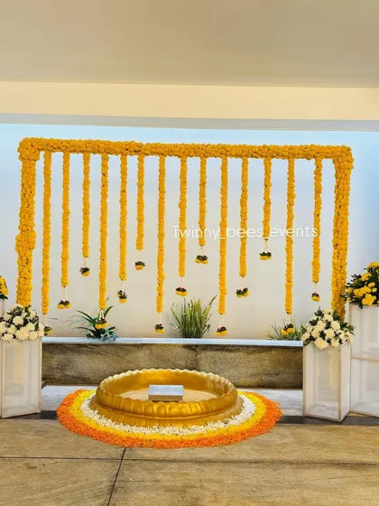 This is another one of our popular Haldi setups. We use a large brass Urli placed on a bed of orange and yellow marigold petals, with a simple marigold arch against a white curtain backdrop.