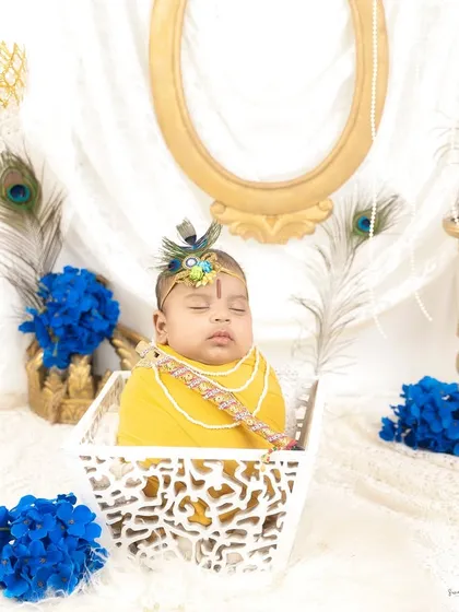 A serene portrait of a baby dressed as little Krishna, sleeping peacefully in a white basket. The setup includes peacock feathers and royal blue flowers for a divine touch.