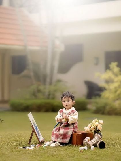A toddler sits with a small easel and paints in a green lawn, with a beautiful house in the background.
