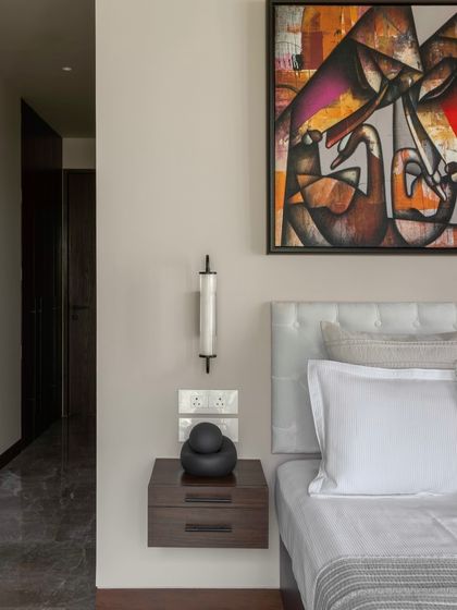 A floating nightstand and a simple wall sconce maintain the minimalist aesthetic in this bedroom at the Parishram residence. The focus remains on the textures of the bedding and the striking artwork.