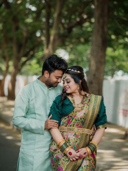 A classic portrait of the couple standing together under a canopy of trees. Their elegant ethnic wear and happy expressions make this a perfect engagement photo.