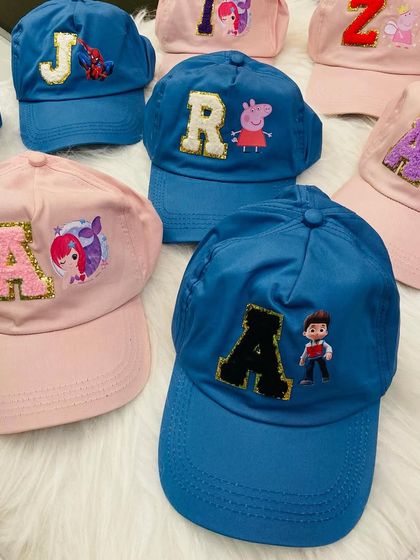 A collection of custom initial caps made as return favors for a kids' party. The image displays a mix of blue and pink caps with patches of characters like Peppa Pig, Paw Patrol, and Spiderman.