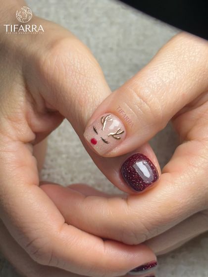 A cute, hand-drawn reindeer face on a nude base, paired with a rich, sparkling burgundy nail for a cozy Christmas feel.