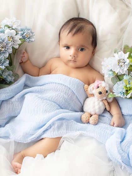 An awake newborn lies on a soft bed of white tulle, wrapped in a blue blanket and surrounded by blue flowers.