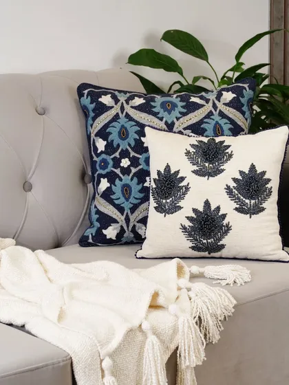 A close-up of elegantly embroidered blue and white pillows on a chic grey sofa. Paired with a soft throw blanket, this image highlights the intricate details and quality of the textiles.