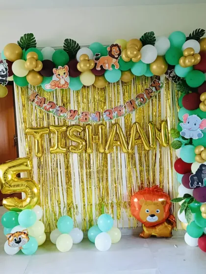 A jungle-themed decoration for Tishaan's 5th birthday, featuring a gold tinsel curtain, a balloon arch with tropical leaves, and cute animal characters.
