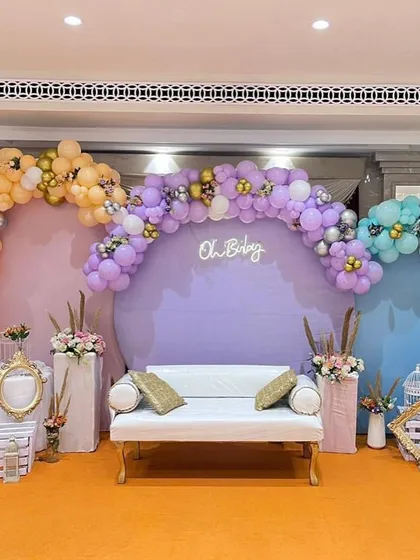 This large-scale baby shower decoration shows how we can transform a venue space. The setup includes three distinct pastel-colored backdrops with balloon arches and a central sofa, providing ample, beautiful seating for the guests of honor.