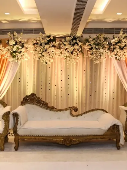An elegant reception stage with a white and gold color scheme. The royal diwan and chairs are set against a backdrop of white flowers and soft, warm lighting.