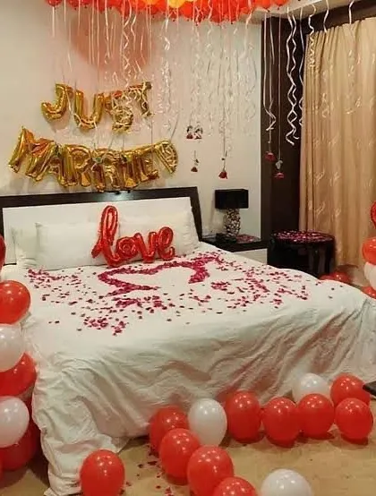 A classic and romantic 'Just Married' room decoration. This setup features red and white balloons, 'Just Married' foil letters, and a heart made of fresh rose petals on the bed, perfect for a memorable first night.