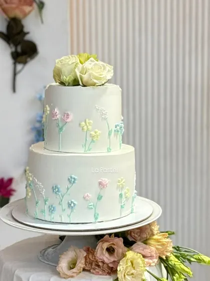 A full view of the two-tier minimal cake with delicate pastel wildflowers piped onto a smooth white canvas, topped with fresh white roses.