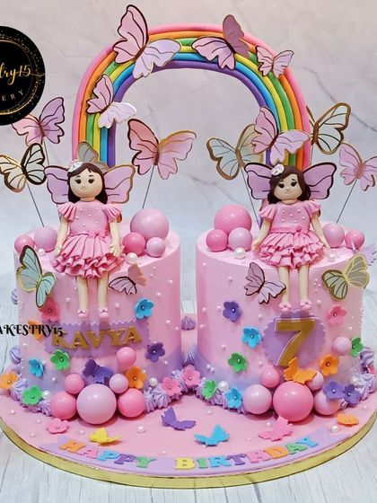 A unique cake design for a 7th birthday, featuring two separate cakes connected by a fondant rainbow. Each cake is topped with a fondant doll and decorated with flowers and butterflies.