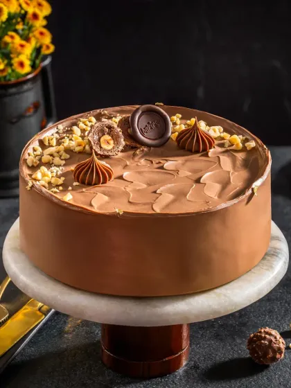 A Ferrero Rocher inspired chocolate cake on a marble stand, styled with whole hazelnuts and chocolate pieces to highlight its ingredients.
