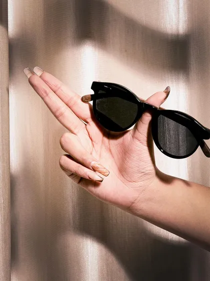 A hand holding a pair of black sunglasses, shot with soft, warm lighting. The pose is elegant and the mood is sophisticated, perfect for a classic eyewear brand.