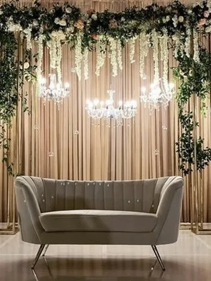 A chic and modern engagement stage setup. The design features a backdrop of cascading florals, elegant chandeliers, and a stylish grey sofa, perfect for a sophisticated celebration.