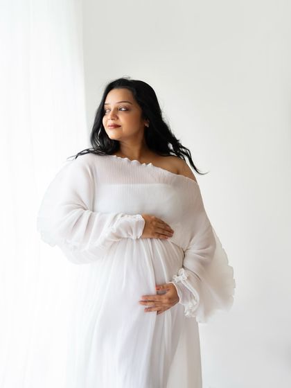 This stunning mom-to-be looks radiant in a flowing white off-the-shoulder gown. The soft fabric and bright, clean background create a classic and angelic maternity portrait.