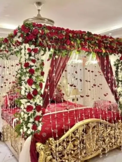 I create luxurious and romantic first night room decorations. This opulent setup features a four-poster bed draped in red fabric and completely covered in fresh red and white roses, with delicate floral strings hanging down for a magical feel.