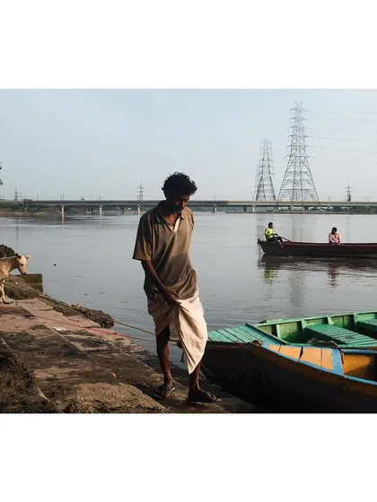 A quiet moment on the banks of the Yamuna, with a boatman standing by his boats and a dog walking along the ghat. In the background, another boat drifts by, completing the tranquil scene.