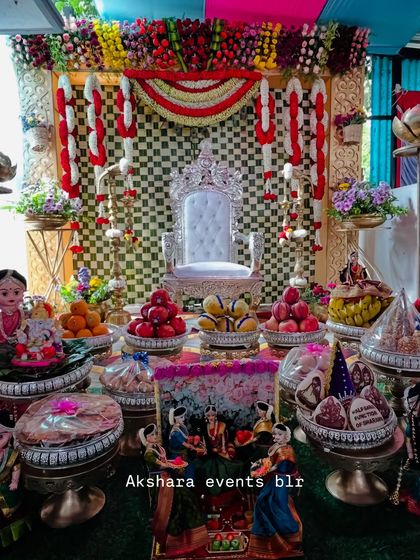 A grand Seemantham setup featuring a silver throne and a checkerboard backdrop in red, yellow, and white. The stage is filled with an elaborate display of traditional offerings and dolls.