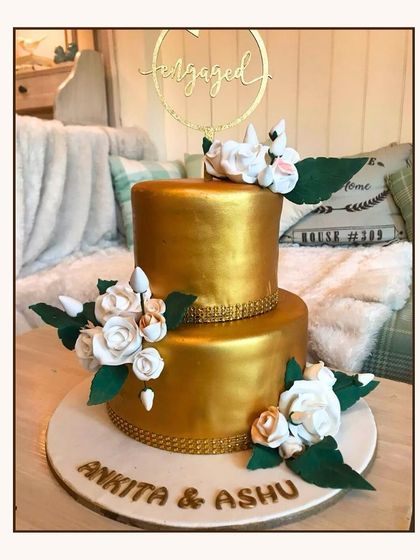 A glamorous two-tier all-gold engagement cake, decorated with elegant white sugar roses.
