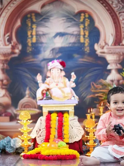 This little boy, dressed in a kurta, smiles brightly while sitting in our detailed Ganpati setup, complete with offerings and traditional lamps.