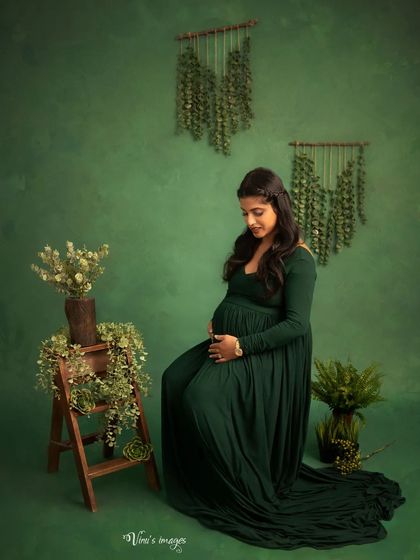 A moment of quiet connection. The mother-to-be sits peacefully, cradling her bump, surrounded by lush greenery that complements her beautiful dark green gown.