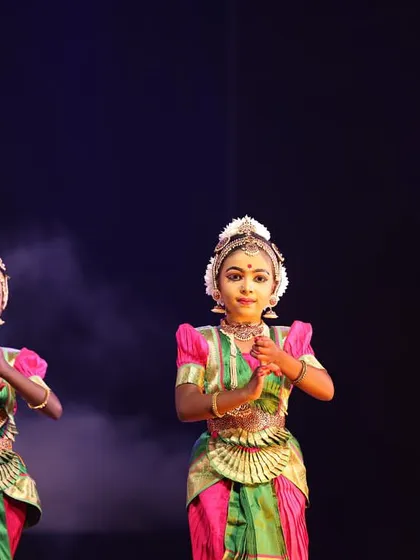 Two young dancers in pink and green costumes, their hands held in a prayerful mudra, their faces reflecting the innocence and devotion of young artists.
