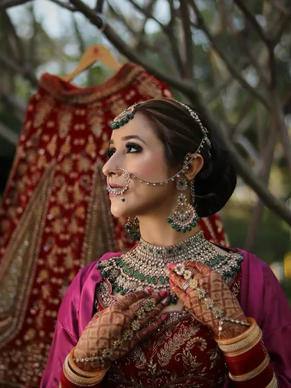 A profile shot capturing the elegance of a bride on her wedding day. This look highlights a sharp contour, a classic bridal bun, and intricate jewelry. The makeup is designed to be long lasting and picture perfect from every angle, with her bridal lehenga hanging in the background, ready for the ceremony.