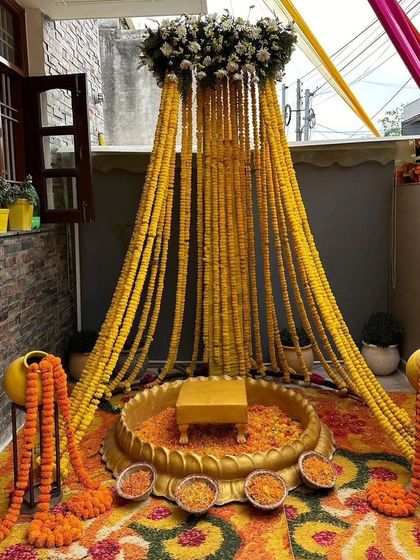 A beautiful canopy-style Haldi decoration for a home's terrace or balcony. The setup uses strings of marigolds hanging from a floral canopy over a traditional brass urli.