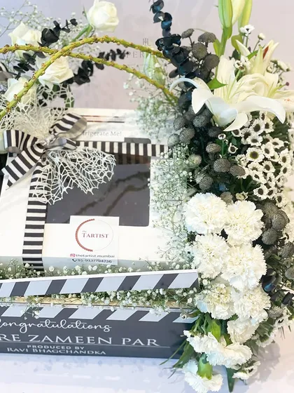 A close-up view of the 'Sitaare Zameen Par' hamper, highlighting the fresh white carnations and lilies arranged around the custom box. The black and white striped ribbon adds a classic, cinematic touch.
