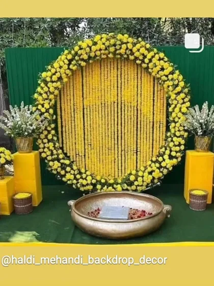 A classic Haldi backdrop for outdoor functions. This setup features a large circular frame filled with strings of yellow marigolds, a green panel background, and a traditional brass urli in the center for the ceremony.