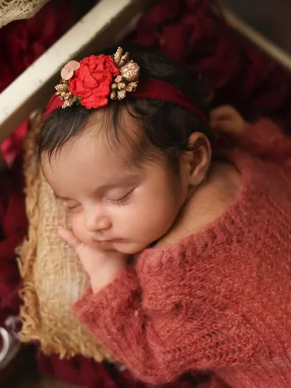 A close-up of a newborn girl in a rich, red-themed setup. The color, flowers, and vintage radio create a warm and romantic atmosphere.