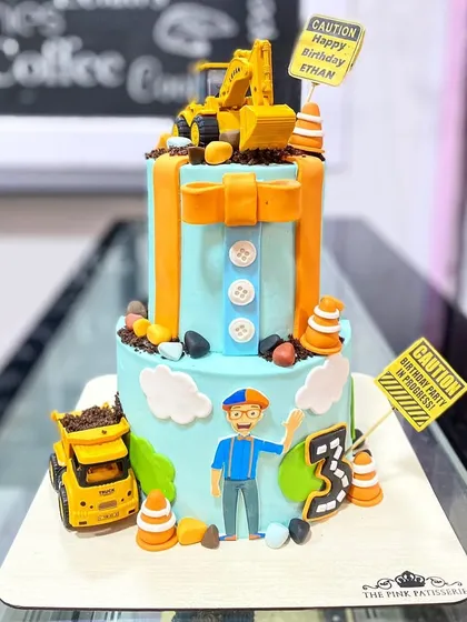 A fun and busy construction-themed cake featuring the character Blippi. This two-tiered cake is decorated with fondant trucks, traffic cones, and edible dirt.