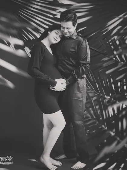 A quiet, heartfelt moment between parents-to-be. The creative use of shadow play with palm leaves adds an artistic and modern touch to this black and white studio portrait.