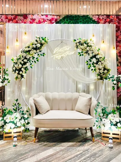 For a sophisticated and timeless celebration, we designed this beautiful white-themed Roka setup. It features a circular arch with white and pink floral arrangements, a 'Better Together' neon sign, and elegant lighting against a backdrop of red and green flower walls.