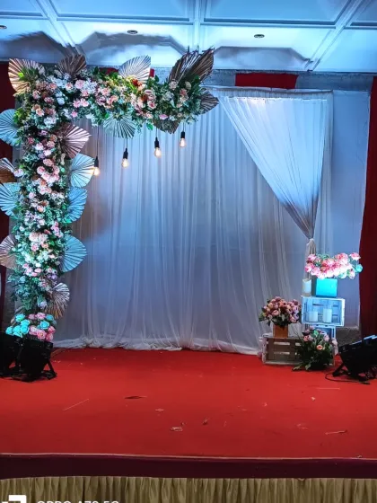 An asymmetrical floral arch on a stage. This modern design uses a mix of pastel flowers and dried palm leaves against a simple white curtain backdrop.