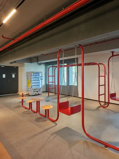 A unique seating installation made from continuous red metal tubing that forms swings and benches. This playful design element is located next to a vending machine for a complete break-time setup.